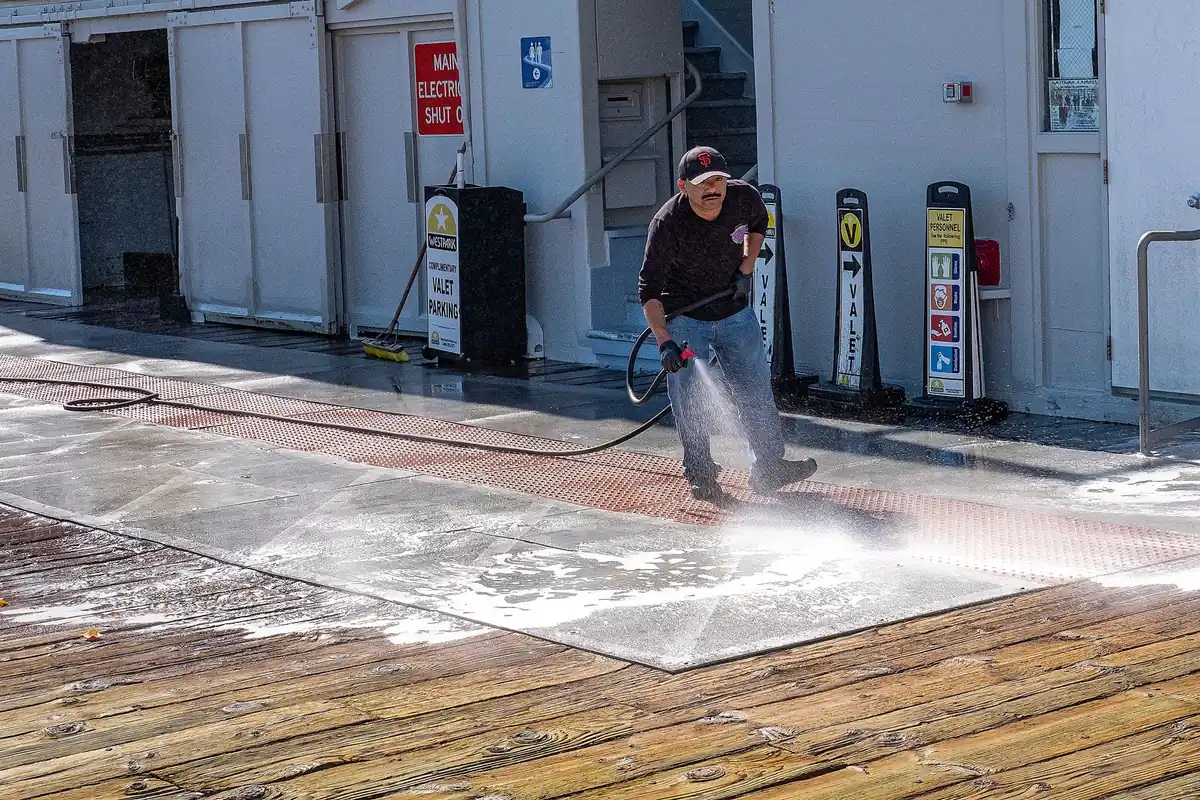 A worker pressure washing boards, a clean strip running beside the gray, dirty surface
