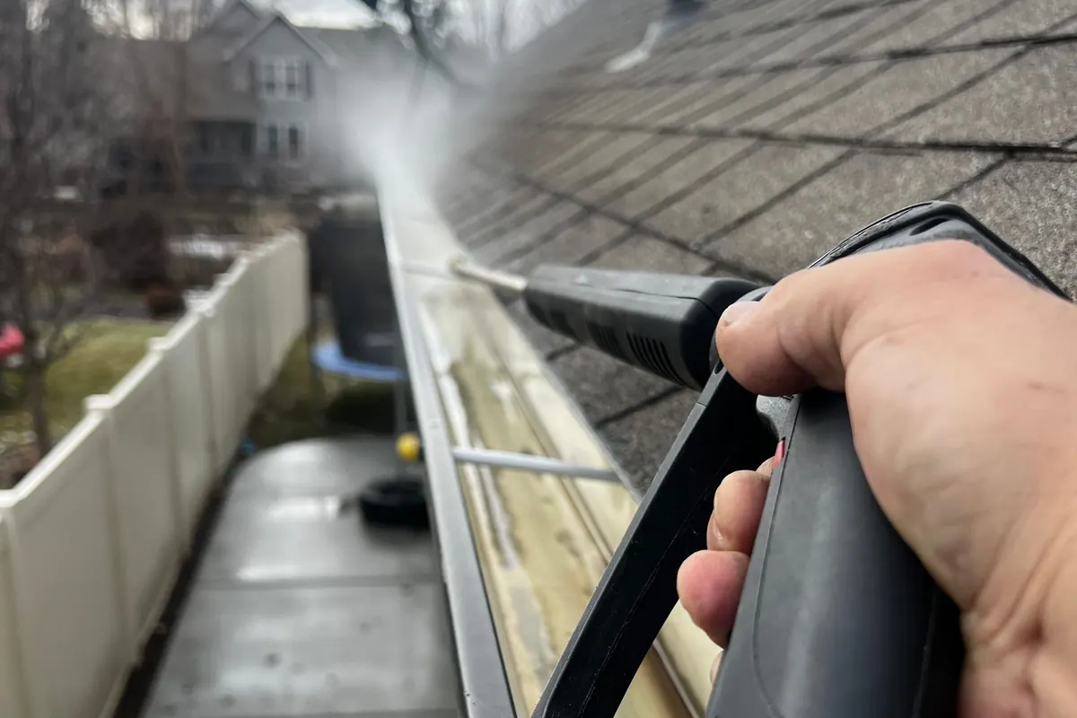 A hand on a pressure washer wand spraying out a gutter along a roof edge