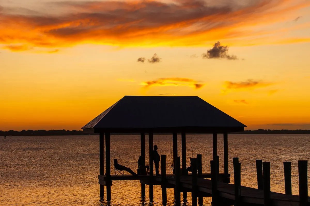 A dock with a covered boat lift silhouetted against an orange sunset over the water