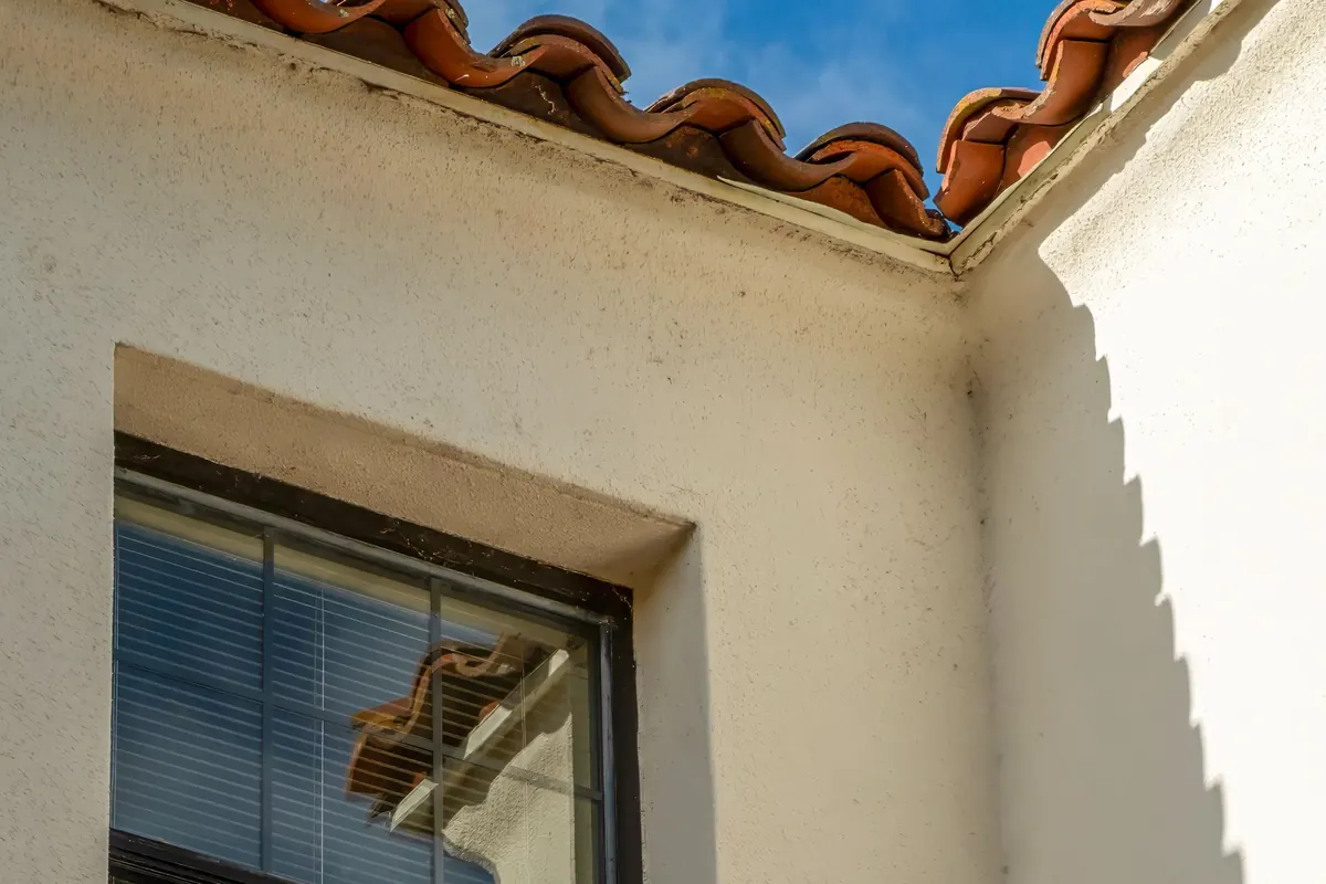 A clean cream stucco wall and window under the edge of a terracotta tile roof